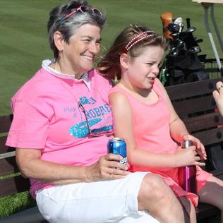 A woman in a pink golf shirt and holding a blue can sits and smiles beside a young girl with a water bottle
